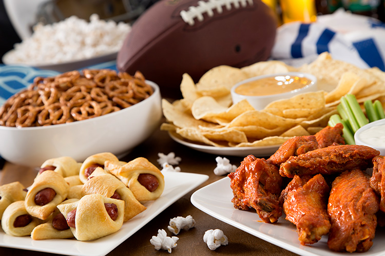 An assortment of tailgating foods spread out on a table. There are buffalo wings, chips and dips, pretzels, pigs in a blanket, and more.